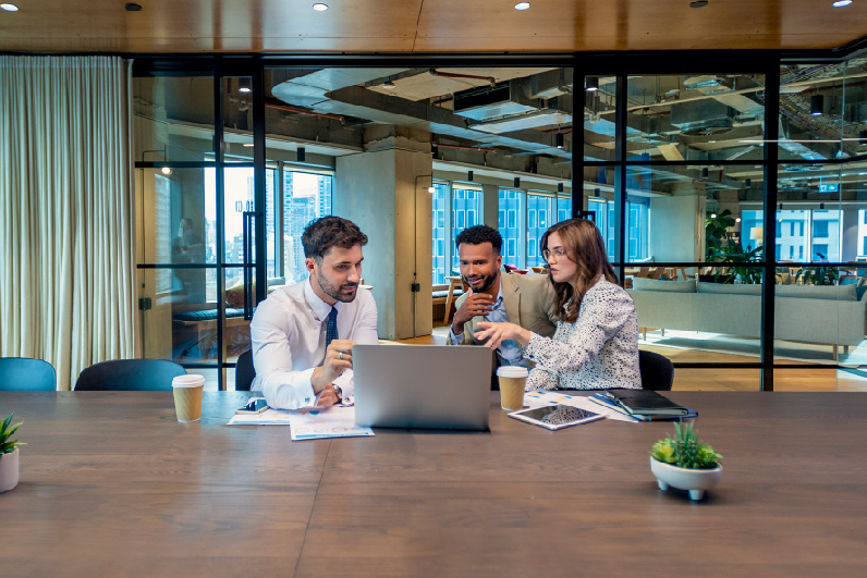 Two men and a woman sit at a large wooden conference table and use a laptop computer.